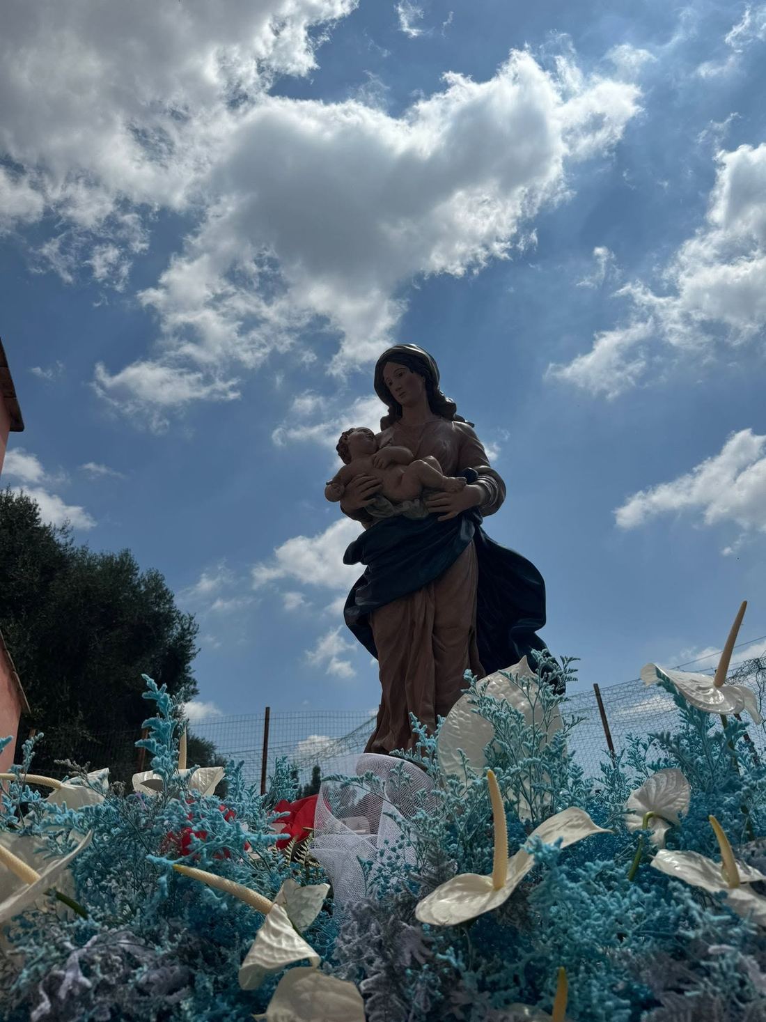 La statua della Madonna con il Bambino, circondata da fiori, sotto un cielo nuvoloso.