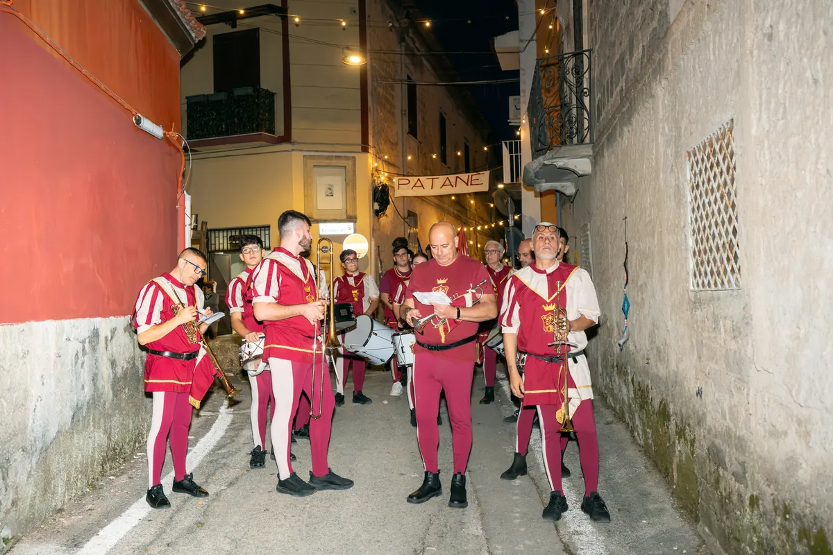 Il corteo in costume rosso e bianco nel vicolo illuminato, sotto lo striscione di Patanè.